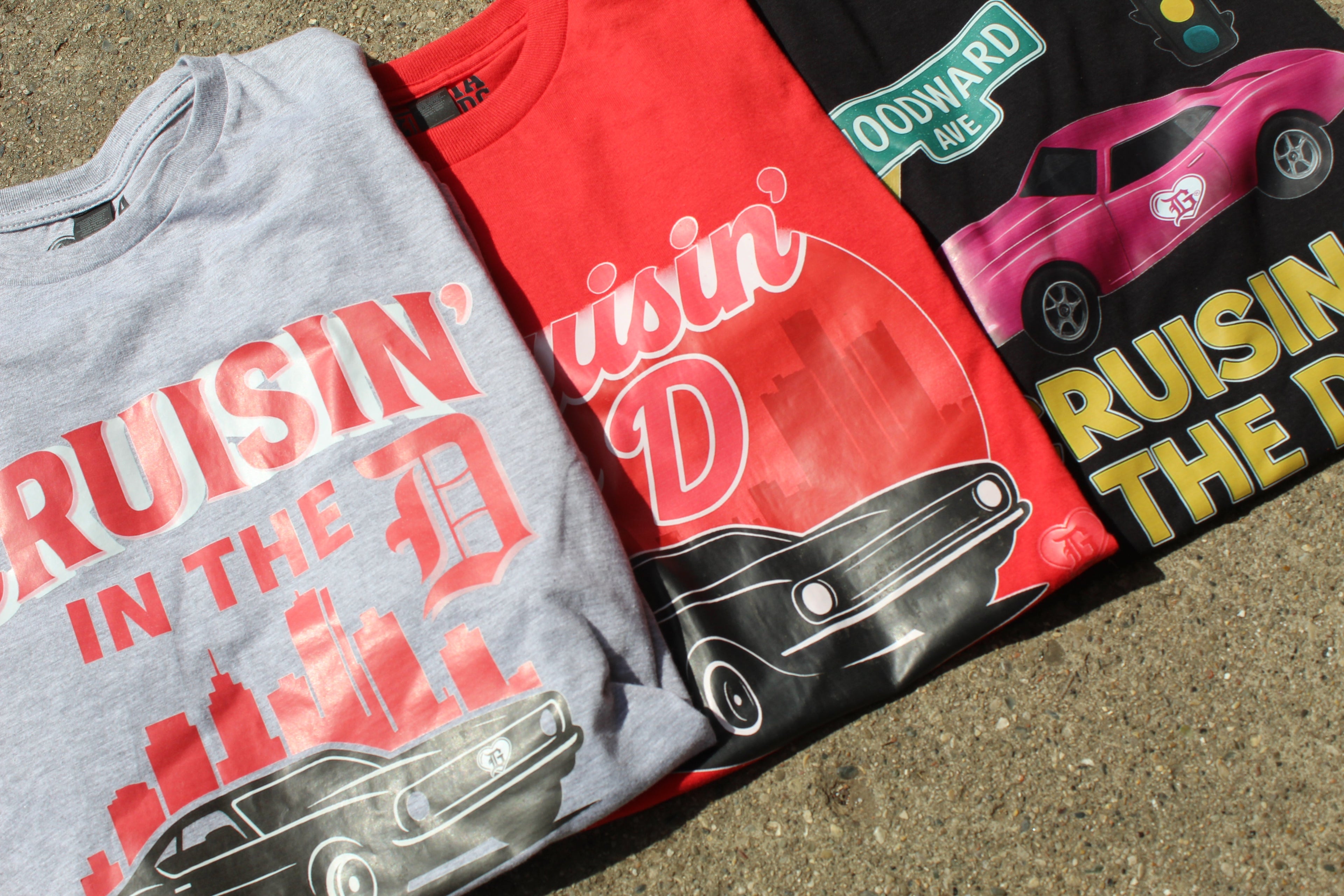 Three “Cruisin’ in the D” T-shirts in grey, red, and black, featuring Detroit skyline and muscle car graphics, by I’m a Detroit Girl Clothing.