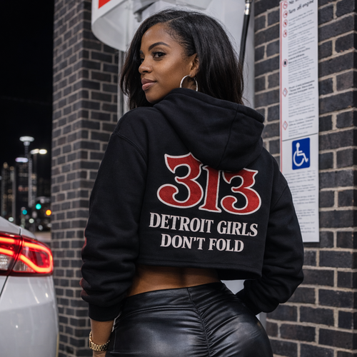 Person wearing a black hoodie with '313 Detroit Girls Don't Fold' text, standing in front of a brick wall.