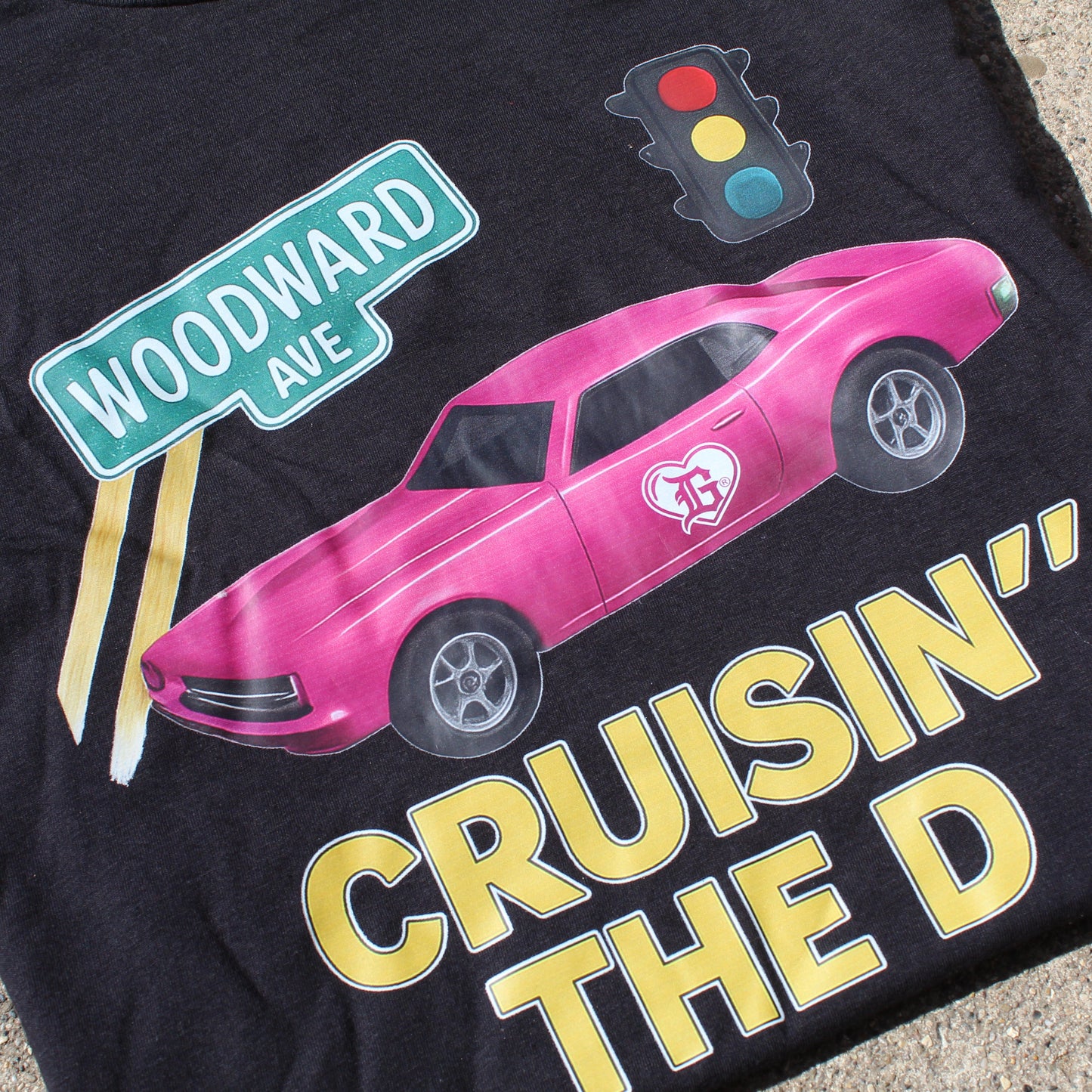 Black t-shirt with pink car graphic, 'Woodward Ave' sign, and 'Cruisin' The D' text on a concrete surface.