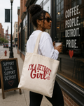A woman walking on a city sidewalk carries a cream canvas tote bag with red text that reads 'I'M A DETROIT Girl' in a storefront setting. The background shows a street scene, a window with white text, and a chalkboard sign.