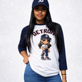 Person wearing a white and navy baseball shirt with a cartoon character, blue jeans, and a navy cap with a logo on a light gray background.