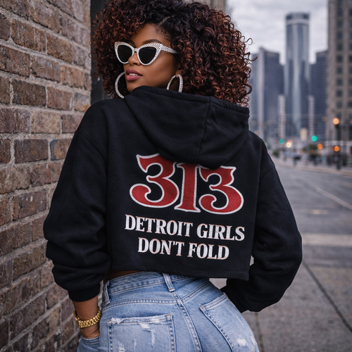 Person wearing a black hoodie with '313 Detroit Girls Don't Fold' text, standing against a cityscape.