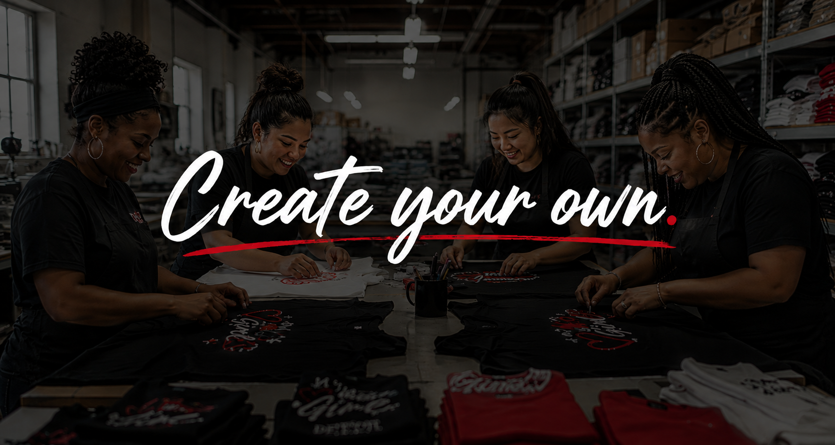 Four women working together in a dark warehouse or studio space, folding and printing black t-shirts. Large white script text across the center reads 'Create your own.