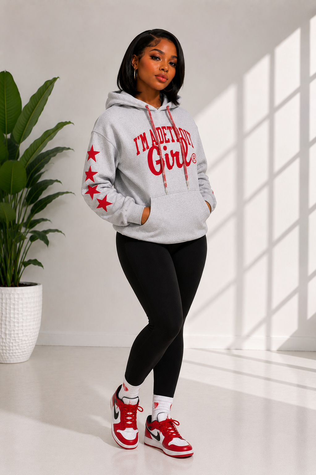 Woman wearing a hoodie with text and red stars, standing in a bright room with a plant.