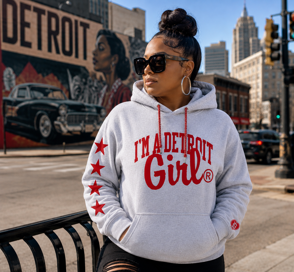 A woman wearing a light gray hoodie with red text reading 'I'M A DETROIT Girl' and red stars on the sleeve, standing on a city street with a Detroit mural in the background.