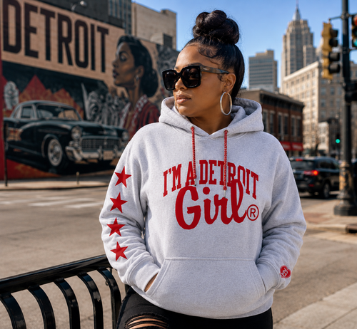 A woman wearing a light gray hoodie with red text reading 'I'M A DETROIT Girl' and red stars on the sleeve, standing on a city street with a Detroit mural in the background.