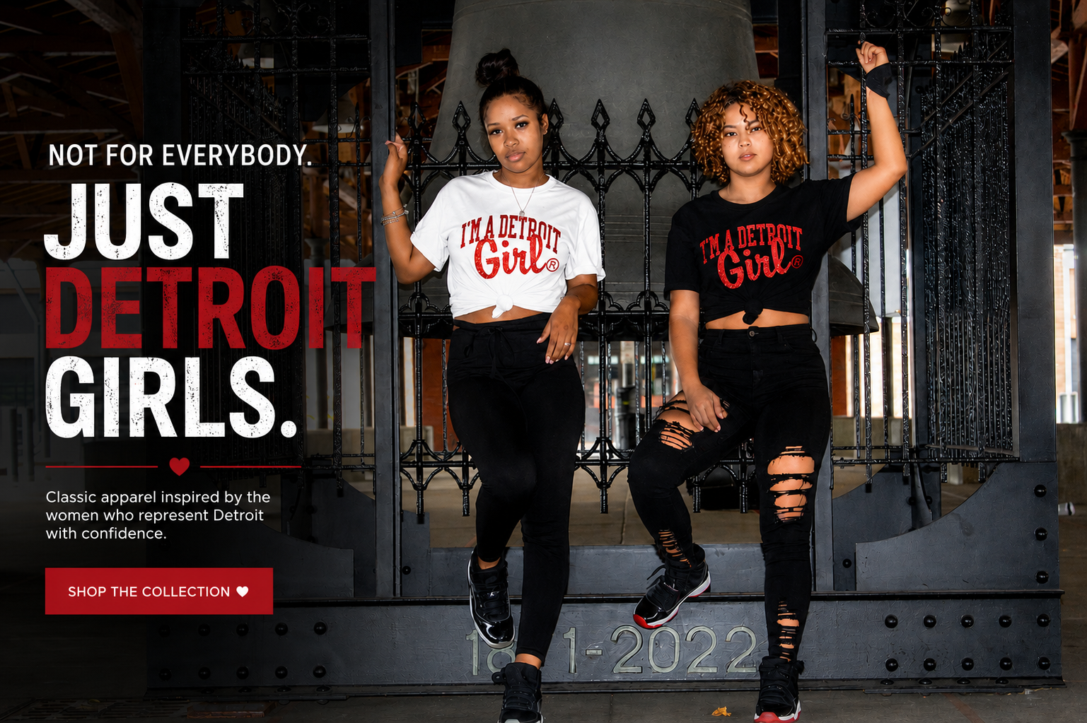 Two women posing in front of a large industrial gate, wearing matching Detroit-themed apparel. One wears a white crop top with red text reading 'I'M A DETROIT Girl' with black leggings, and the other wears a black crop top with red text reading 'I'M A DETROIT Girl' with black ripped leggings.