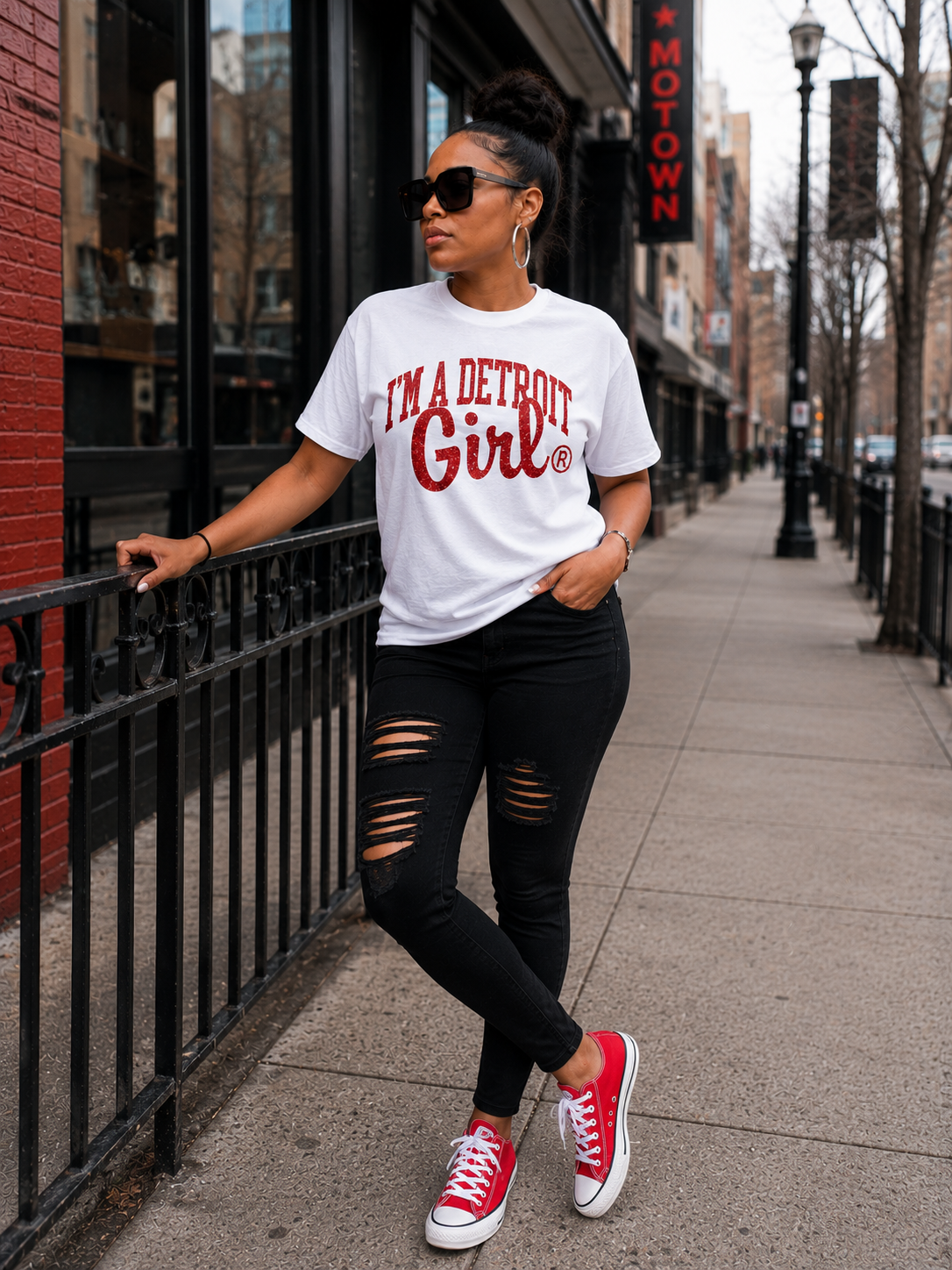 A woman stands on a city sidewalk wearing a white graphic T-shirt that reads 'I'M A DETROIT Girl' in red, black ripped skinny jeans, and red low-top sneakers. She is leaning against a black metal railing next to a storefront.